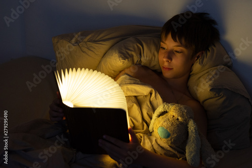 Adorable boy reads book before sleeping in bed at home with his old toy. Happy childhood.