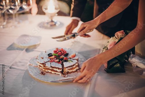 Stylish happy bride and groom cutting together wedding cake with fruits at wedding reception outdoors in the evening. Couple hands cutting cake in soft candle light in night.