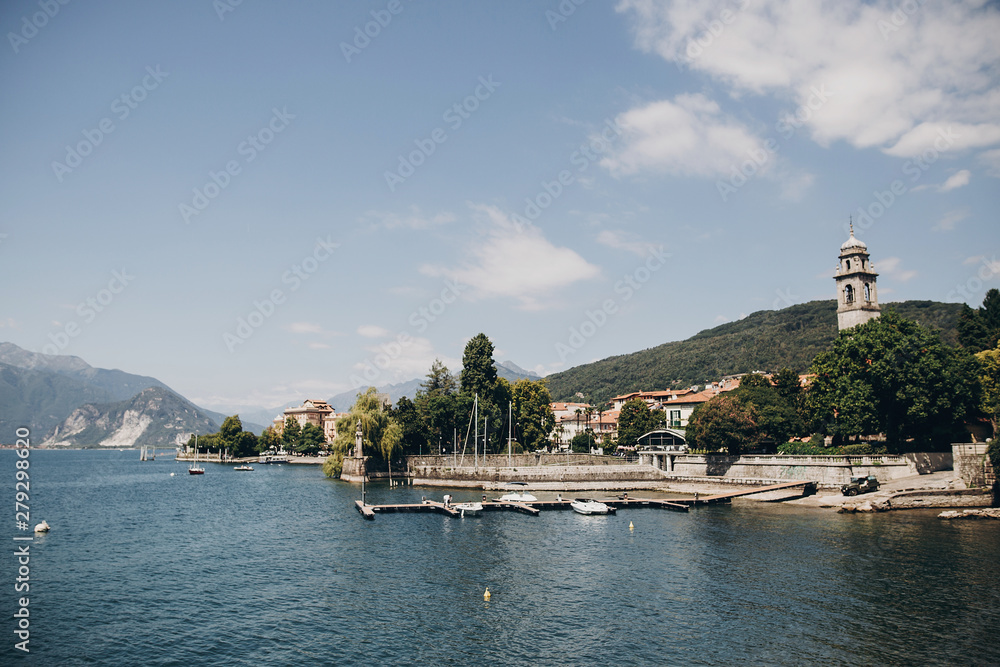 Beautiful view on old buildings and boats on lake in Stresa city, Italy. Architecture and shore on Lago Maggiore in sunny day on background of palms and mountains. Summer vacation in Europe Beautiful view on old buildings and boats on lake in Stresa city, Italy. Architecture and shore on Lago Maggiore in sunny day on background of palms and mountains. Summer vacation in Europe
