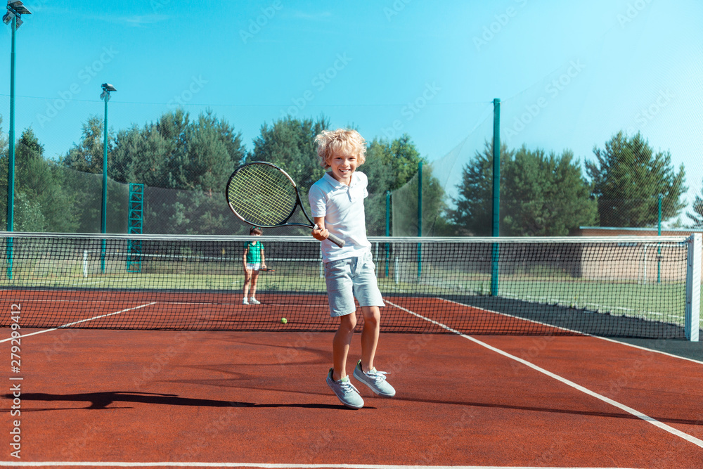 Cheerful curly boy feeling happy while playing tennis outside