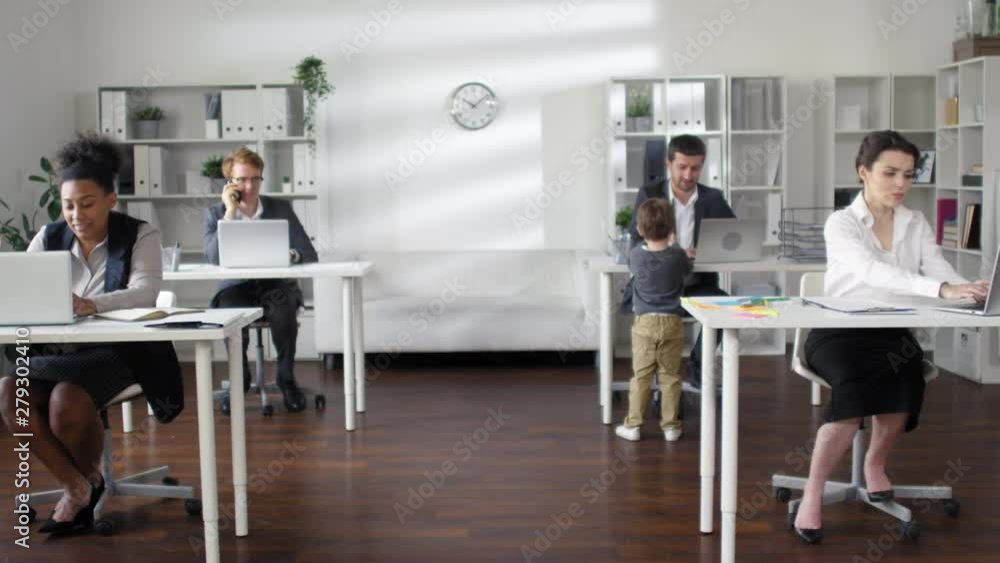 Cute little boy playing with sticky notes while multiethnic business people sitting at their desks and working Cute little boy playing with sticky notes while multiethnic business people sitting at their desks and working