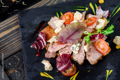 Beautiful serving of toast with meat, tomato, cheese and fresh herbs on a dark plate on a rustic wooden tray