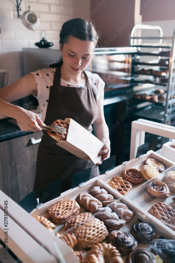 Pretty smiling female worker selecting muffins from a counter display in small bakery to sell to a customer, Family business concept