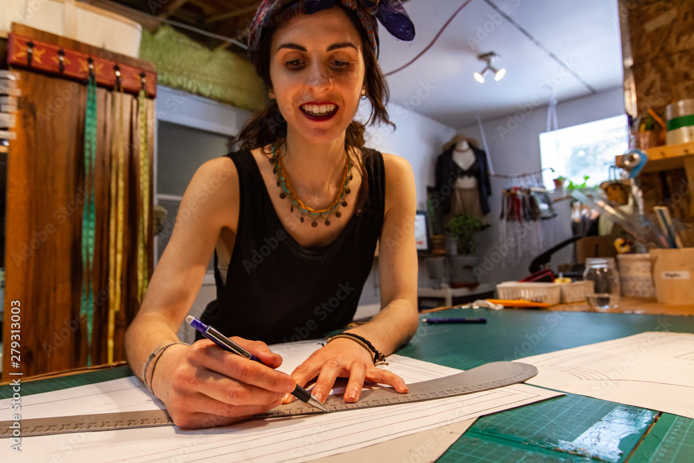 Fototapeta premium Fashion design on a grid sketching table. A close up view of a female tailor using ruler and technical pen to plan new clothing, she looks happy with the design process.