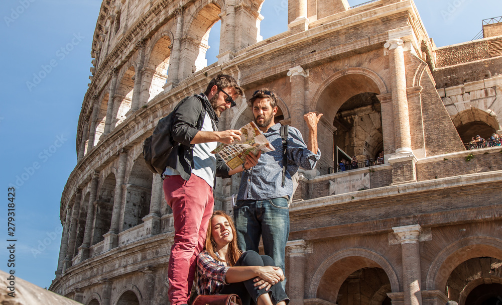 Three happy young friends tourists standing and sitting in front of colosseum in rome reading city map guide