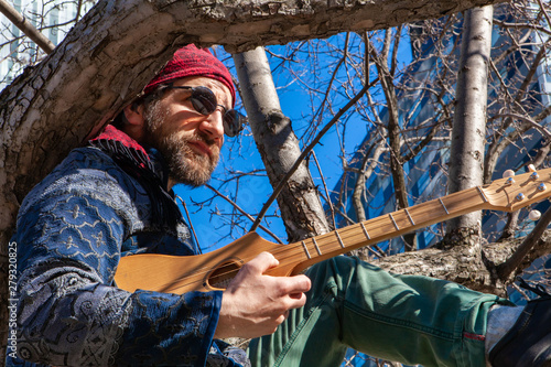 Creative man sits in tree by skyscraper. A close-up view of a spiritual person sitting on a tree branch downtown, holding a wooden string instrument, seeking inspiration in city surroundings. © Valmedia