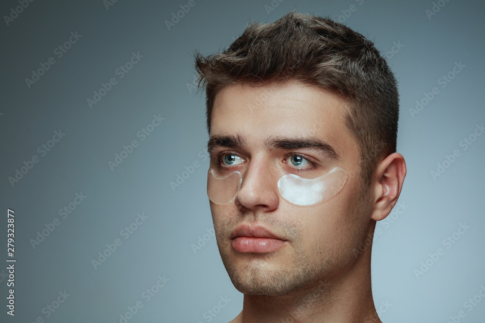 Naklejka premium Close-up profile portrait of young man isolated on grey studio background. Male face with collagen patches under eyes. Concept of men's health and beauty, cosmetology, body and skin care. Anti-aging.