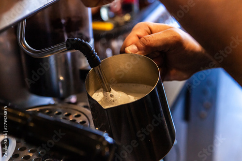 Barista froths milk in coffeehouse. A closeup view on the working hands of a coffeshop server, using a frother to create microfoam from warm milk, to be added into a cappuccino in a bistro.