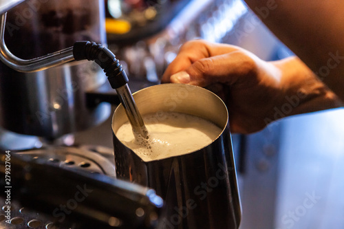 Barista froths milk in coffeehouse. A closeup view on a person holding a jug of milk under the steam baffle of a coffee machine  used to heat and aerate milk for latte art in a coffee shop.