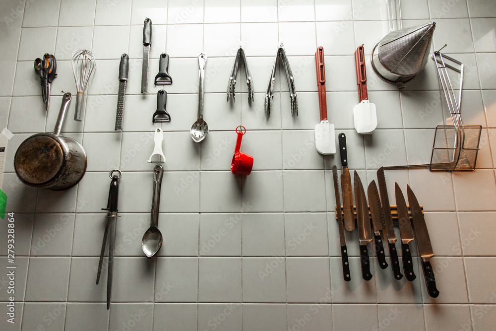 Kitchen utensils hanging on wall. A closeup view of cooking utensils  hanging on a tiled wall inside a restaurant kitchen, professional tools are  neatly organized for easy access Photos