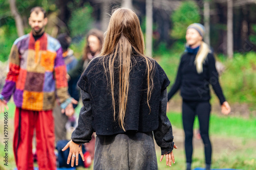 Fusion of cultural & modern music event. A girl with long blonde hair is seen from the rear, wearing native clothes and standing among a group of people performing spiritual meditation outdoors.