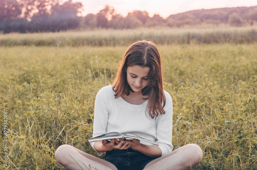Christian teenage girl holds book in her hands. Reading the book in a field during beautiful sunset. The girl sitting on a grass, reading a book. Rest and reading Christian teenage girl holds book in her hands. Reading the book in a field during beautiful sunset. The girl sitting on a grass, reading a book. Rest and reading