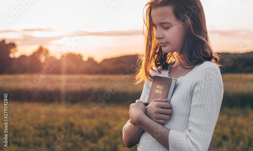 Christian teenage girl holds bible in her hands. Reading the Holy Bible in a field during beautiful sunset. Concept for faith, spirituality and religion. Peace, hope photo