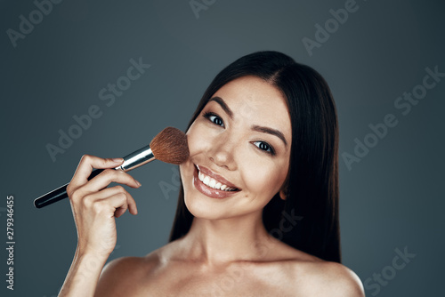 Putting on some make up. Beautiful young Asian woman looking at camera and applying blush brush while standing against grey background