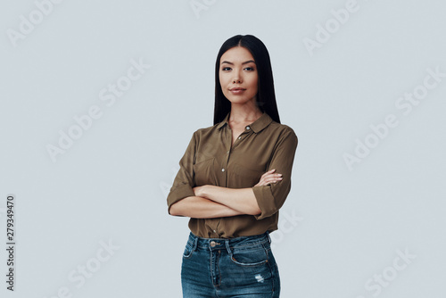 Young and confident. Attractive young Asian woman looking at camera and keeping arms crossed while standing against grey background