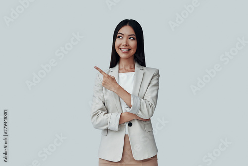 And here comes... Attractive young Asian woman looking away and pointing copy space while standing against grey background
