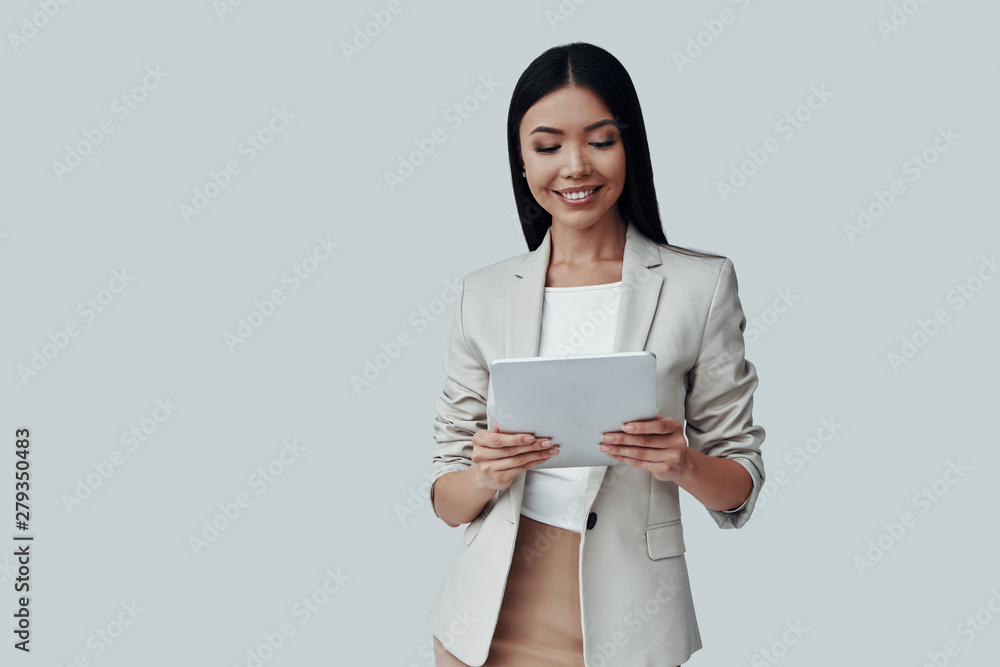 Confident expert. Attractive young Asian woman using digital tablet and smiling while standing against grey background Confident expert. Attractive young Asian woman using digital tablet and smiling while standing against grey background