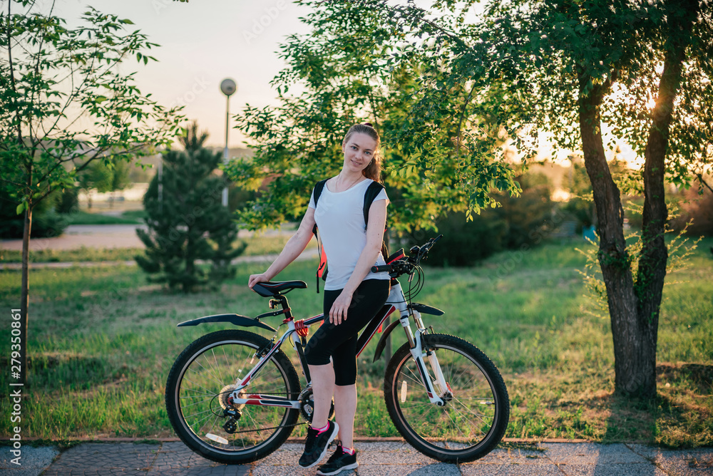 Active life. Sporty young blonde girl in sports clothes with a backpack rides a bike and smiles at the camera in a green Park on a summer evening at sunset. Sport and healthy lifestyle