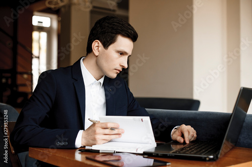 Side view portrait of a confident seriously young manager working in a modern office writing ina notebook and looking at a laptop.
