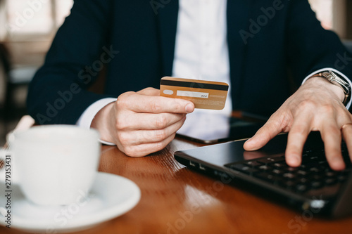 Close up of a male hand dressed in suit holding a gold credit card while sitting on a table with a laptop.