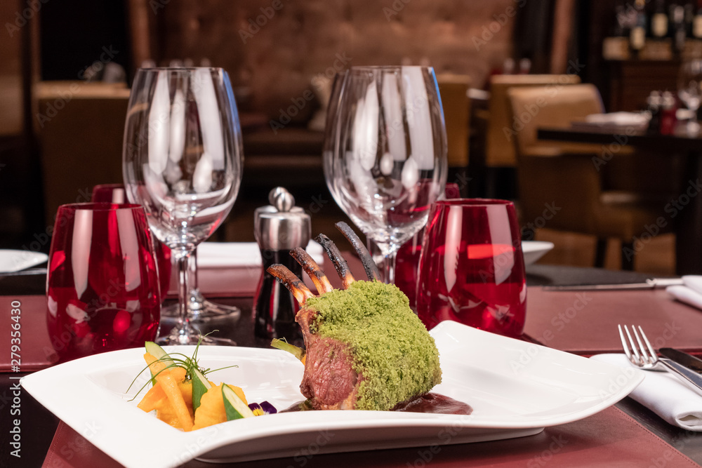 Piece of lamb rack with bone roasted with green herb, bread, cheese crumbs cover, served with sweet potatoes puree and baby vegetables, red wine sauce, in a restaurant elegant setting, on white plate Piece of lamb rack with bone roasted with green herb, bread, cheese crumbs cover, served with sweet potatoes puree and baby vegetables, red wine sauce, in a restaurant elegant setting, on white plate