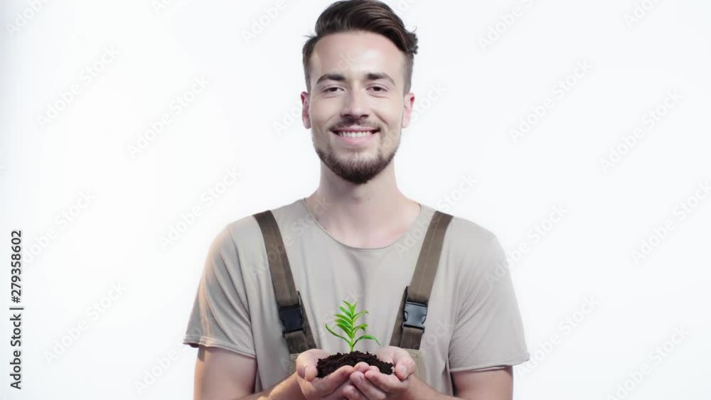 front view of handsome gardener holding handful of soil with green sprout, closing eyes and blinking while smiling at camera