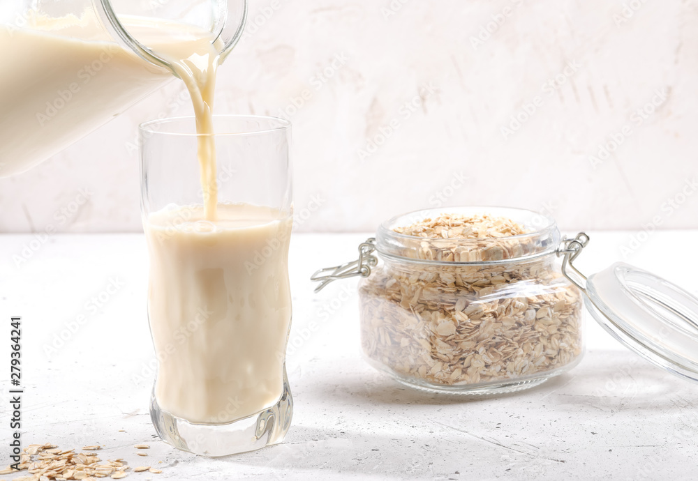 Oat milk pouring from glass decanter into drinking glass and glass jar with oat flakes on light background. Oat milk pouring from glass decanter into drinking glass and glass jar with oat flakes on light background.