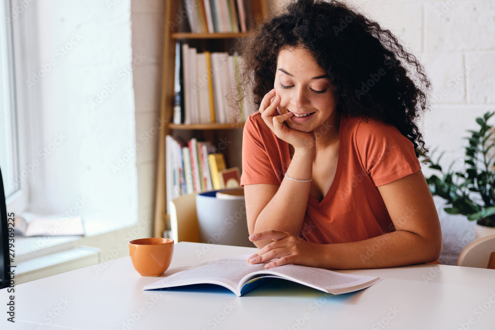 Young beautiful smiling woman with dark curly hair sitting at the table near window with orange cup of coffee dreamily reading book at cozy home Young beautiful smiling woman with dark curly hair sitting at the table near window with orange cup of coffee dreamily reading book at cozy home