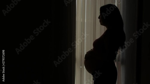 Pregnant beautiful Latin American woman standing looking out at window and caressing her stomach, silhouette