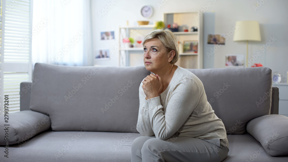 Depressed mature woman praying, sitting on sofa, life difficulties and crisis