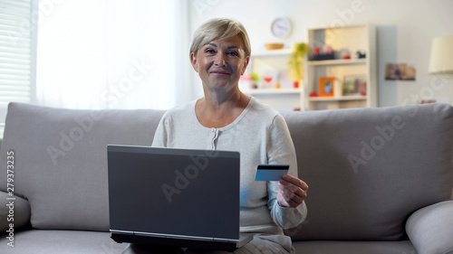 Mature woman using laptop, holding credit card and smiling at camera, banking