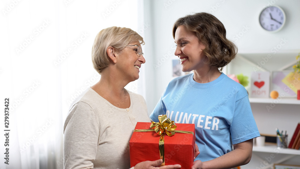 Smiling lady in volunteer t-shirt giving gift box to aged lady, holiday charity Smiling lady in volunteer t-shirt giving gift box to aged lady, holiday charity