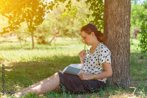 A girl with a notebook under a tree, thoughtfully holding a pencil near the lips.