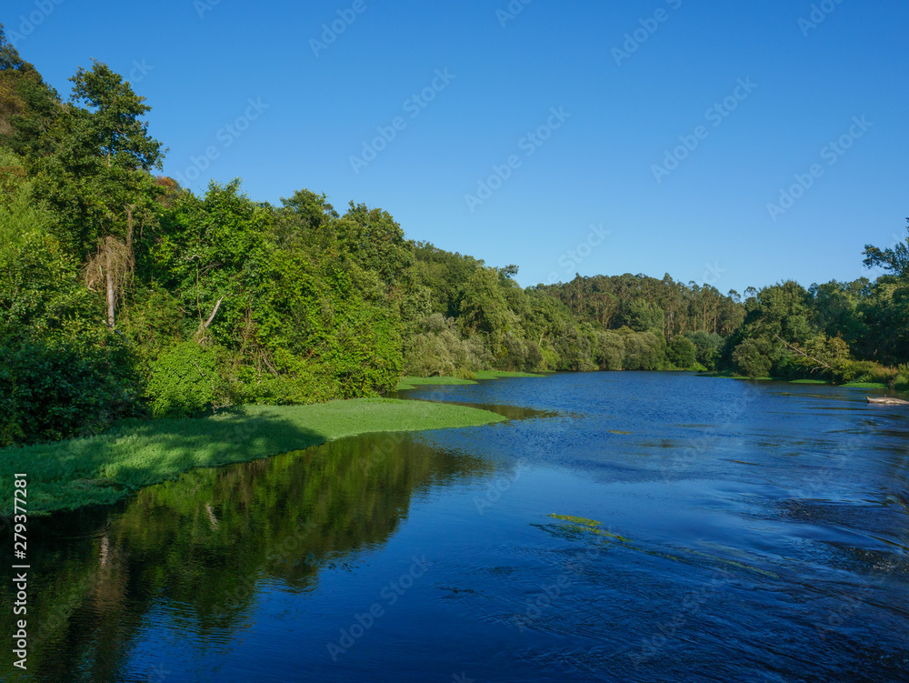 Beautiful summer scene over Ave river in Vila do Conde, Portugal on bright sunny day with blue sky and green trees lining river banks.