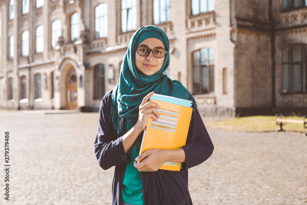 Smiling muslim young female student holding books standing near college. Happy Arabian girl in hijab. Asian woman on training.