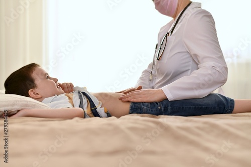 The boy lies on the bed at home, smiles and looks at the doctor who feels his tummy.