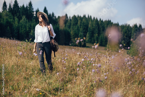 Hipster girl with backpack traveling in sunny mountains, walking in wildflower meadow. Stylish happy woman in hat exploring and hiking on top of mountain. Wanderlust and travel concept