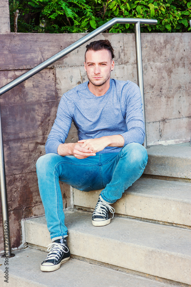 Young Handsome American College Student thinking outside in New York City, wearing gray long sleeve T shirt, blue jeans, black sneakers, sitting on stairs by wall on campus, relaxing, thinking..