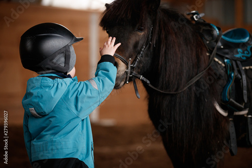Closeup of a kid stroking a horse. The boy is preparing to learn pony riding in the cold season. Hippotherapy for the development of the baby. Child in a warm suit and helmet for safety. Safety first