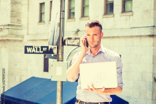 Young handsome man traveling, working in New York City, wearing long sleeve, light color shirt, standing outside office by Wall Street sign under sun, working on laptop computer, talking on cell phone