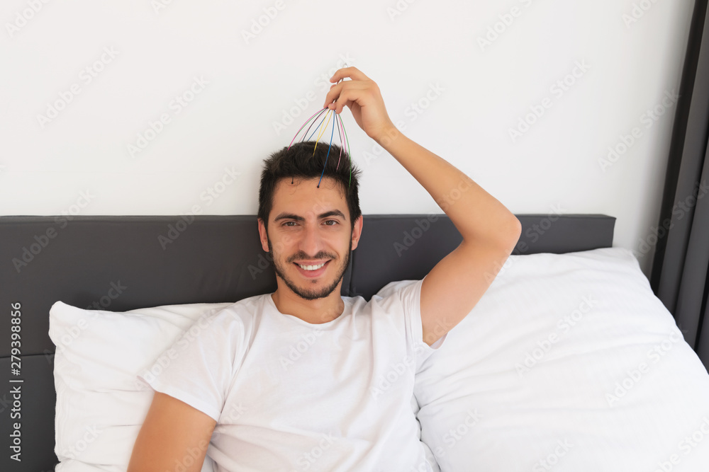 Young handsome man with a beard sits in his bed. He makes himself a head massage with a special hand massager. Young handsome man with a beard sits in his bed. He makes himself a head massage with a special hand massager.