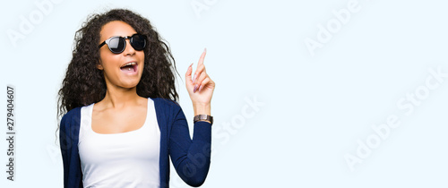 Young beautiful girl with curly hair wearing fashion sunglasses pointing finger up with successful idea. Exited and happy. Number one. © Krakenimages.com