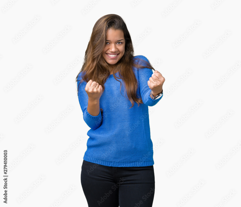 Young beautiful brunette woman wearing blue sweater over isolated background very happy and excited doing winner gesture with arms raised, smiling and screaming for success. Celebration concept. Young beautiful brunette woman wearing blue sweater over isolated background very happy and excited doing winner gesture with arms raised, smiling and screaming for success. Celebration concept.