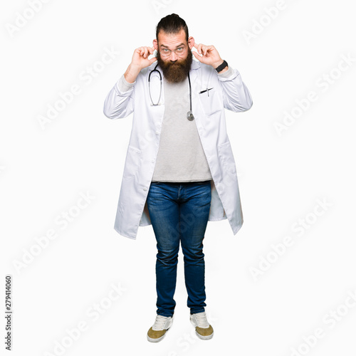 Young blond doctor man with beard wearing medical coat Smiling pulling ears with fingers, funny gesture. Audition problem