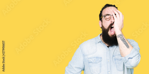 Young hipster man with long hair and beard wearing glasses Yawning tired covering half face, eye and mouth with hand. Face hurts in pain.