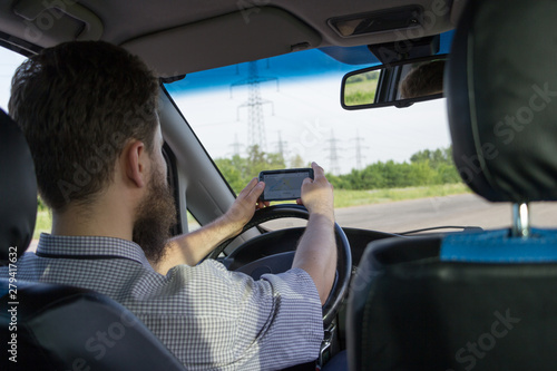 Young bearded man sitting behind the wheel and looking for route by navigation map in smartphone. Rear view.