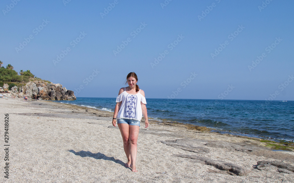 Girl in shorts and white blouse on the beach by the sea.. Red-haired woman on a sandy beach. A holiday in the South