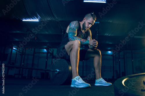 Young healthy man athlete preparing for training in the gym. Single male model sitting on the barbell before the practice. Concept of healthy lifestyle, sport, fitness, bodybuilding, crossfit.