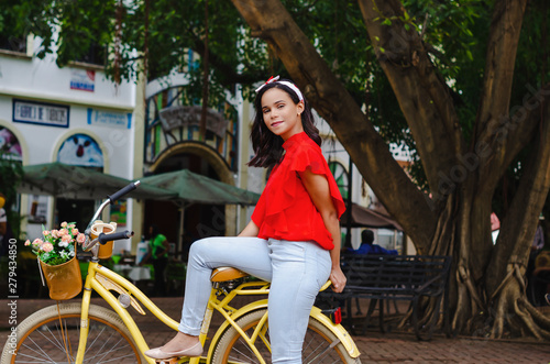 Outdoor portrait of young beautiful girl 19 to 25 years old posing in street. Brunette. riding a retro styled bicycle. Wearing red blouse With a penetrating camera look. City lifestyle.