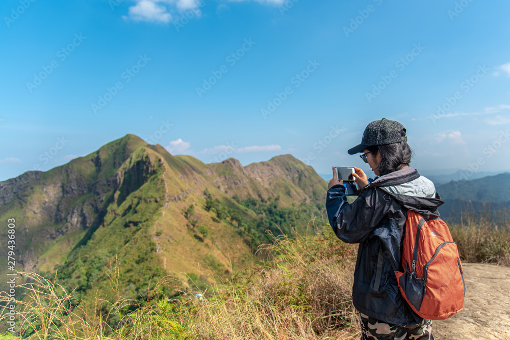young asian woman backpacker,traveling woman standing on top of mountain  and using mobile phone to take photo of nice view.